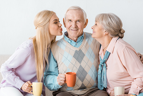 Senior man sitting between younger blonde woman and senior woman both looking at him, all holding coffee cups